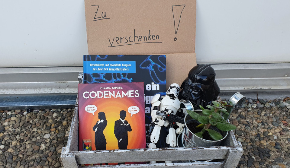 Holzkiste mit Pappschild zu verschenken und Gegenständen Spiel Codenames, weiße Starwarsfigur, eine schwarze Maske und ein Blumentopf mit Pflanze