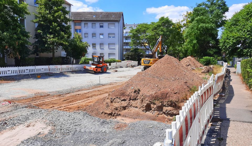 Blick auf eine Straßenbaustelle in der Saarlandstraße. Die Fahrbahn ist großflächig geöffnet und mit einer Schotterschicht vorbereitet. Im Vordergrund liegen mehrere große Erdhaufen, dahinter arbeiten ein Bagger und eine Walze auf der Baustelle. Weiße Absperrbaken begrenzen den Arbeitsbereich. Rechts verläuft ein Gehweg entlang der Baustelle. Im Hintergrund stehen mehrgeschossige Wohnhäuser und große, grüne Bäume bei sonnigem Wetter.