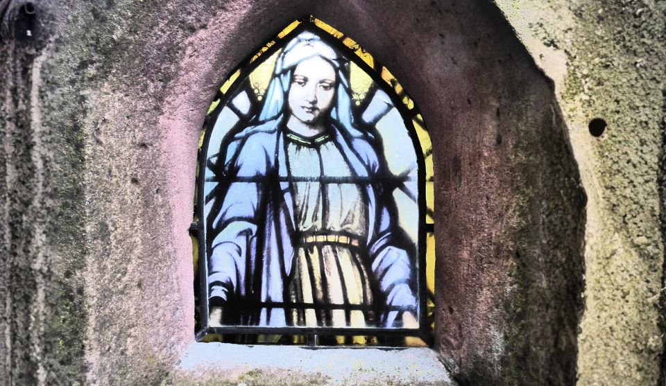 Nahaufnahme eines spitzbogenförmigen Buntglasfensters in einer steinernen Einfassung. Das Fenster zeigt eine weibliche, religiös anmutende Figur in einem hellen Gewand und blauem Umhang.
