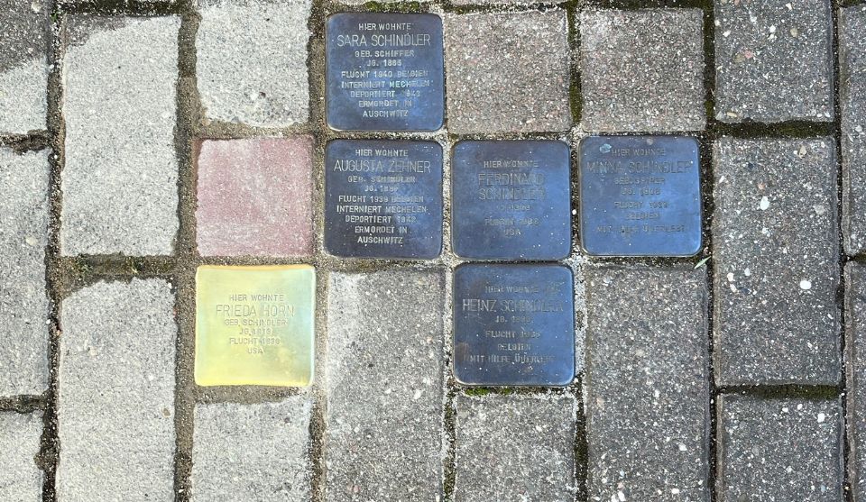 Mehrere Stolpersteine sind zwischen grauen Pflastersteinen in den Boden eingelassen. Die Gedenksteine erinnern mit Namen und Angaben zu Flucht, Deportation und Ermordung an Mitglieder der Familie Schindler. Der linke Stein glänzt und scheint neu zu sein. Er erinnert an Frieda Horn, geborene Schindler.