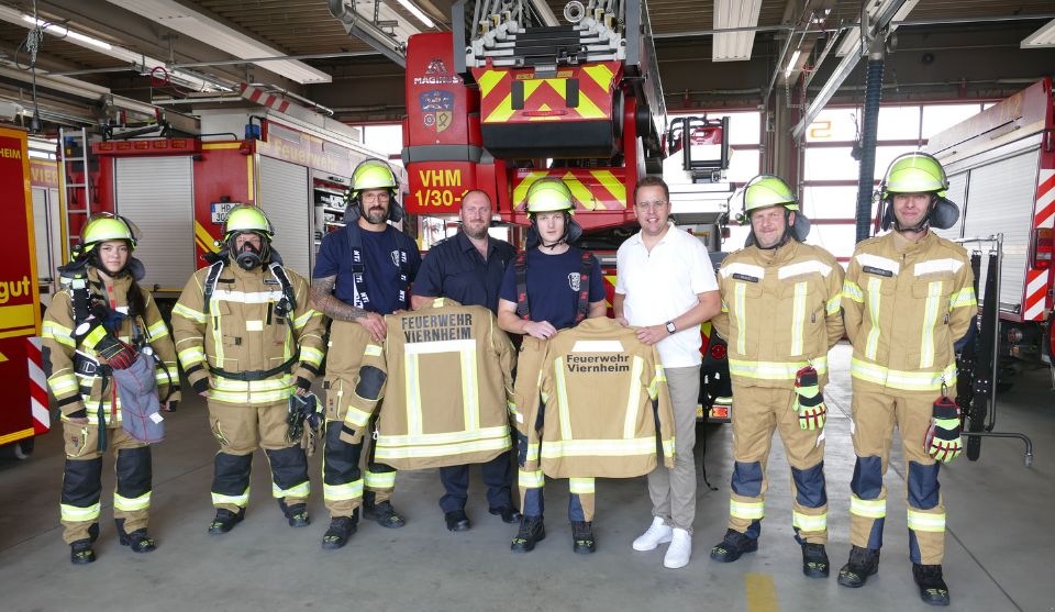 Acht Personen stehen in einer Fahrzeughalle der Feuerwehr vor mehreren Einsatzfahrzeugen. Sechs von ihnen tragen vollständige Feuerwehrschutzkleidung mit Helm, zwei tragen Dienstkleidung beziehungsweise Zivilkleidung. Zwei Feuerwehrangehörige halten neue Einsatzjacken mit der Aufschrift „Feuerwehr Viernheim“ in den Händen. Im Hintergrund sind ein Drehleiterfahrzeug und weitere Löschfahrzeuge zu sehen.