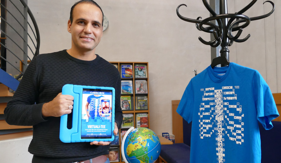 Michael Silva hält in den Händen Curiscope Virtuali-Tee, rechts daneben hängt an einem Kleiderständer ein blaues T-Shirt mit Aufdruck QR Code