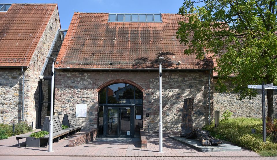 Außenansicht des Eingangs der Stadtbibliothek Viernheim in einem historischen Steingebäude mit rotem Ziegeldach. Vor dem Eingang befinden sich Sitzbänke, Bäume und eine Skulptur.