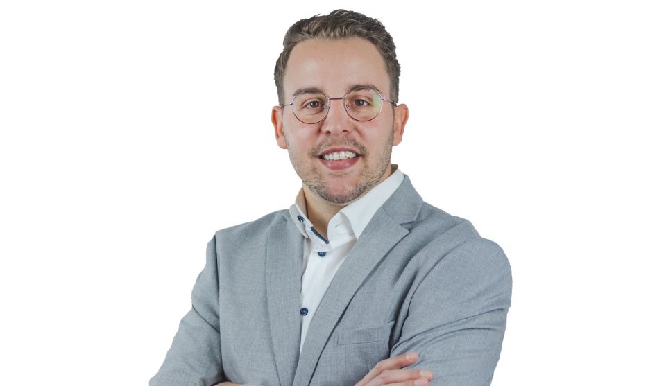 Porträt eines lächelnden Mannes, Jörg Scheidel, mit kurzen dunklen Haaren und runder Brille vor weißem Hintergrund. Er trägt ein weißes Hemd und ein hellgraues Sakko und hat die Arme vor der Brust verschränkt.