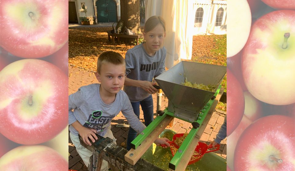 Zwei Kinder helfen beim Keltern von Äpfeln im Museumsgarten. Gemeinsam bedienen sie eine Apfelmühle, in der die Äpfel für die Saftherstellung zerkleinert werden.