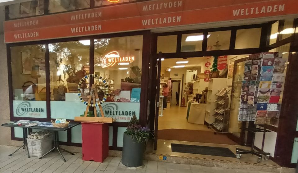 Außenansicht des Weltladens Viernheim mit geöffnetem Eingang und Schaufenster. Vor dem Geschäft stehen ein kleines Glücksrad, ein Tisch mit Informationsmaterial und ein Kartenständer.