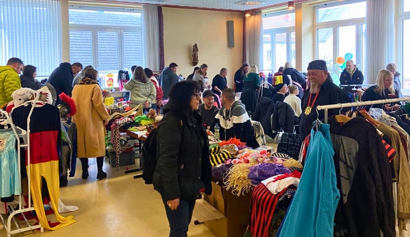 Ein hell beleuchteter Raum voller Menschen, die an verschiedenen Ständen nach bunten Fastnachtskostümen, Hüten und Accessoires stöbern. Kinder- und Erwachsenenverkleidungen hängen an Kleiderständern, auf den Tischen liegen Perücken, Stofftiere und Dekoartikel. Ein Verkaufsbereich ist mit Luftballons geschmückt. Die Stimmung ist lebendig, Besucher unterhalten sich und genießen Getränke und Kuchen. Das Bild vermittelt eine fröhliche, fastnachtliche Atmosphäre.