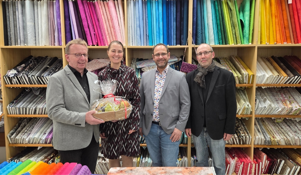 Vier Personen stehen in einem Fachgeschäft für Stoffe vor einem großen Regal, das farbenfrohe Stoffrollen sortiert nach Farben zeigt. Links im Bild hält Bürgermeister Matthias Baaß einen Geschenkkorb mit Zellophanverpackung. Neben ihm stehen Inhaberin Renate Müller, Alexander Schwarz und Claus Bunte von der Wirtschaftsförderung. Alle lächeln in die Kamera. Im Vordergrund liegen weitere bunte Stoffe und Nähzubehör.