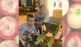 Zwei Kinder helfen beim Keltern von Äpfeln im Museumsgarten. Gemeinsam bedienen sie eine Apfelmühle, in der die Äpfel für die Saftherstellung zerkleinert werden.