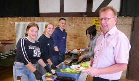 Bürgermeister Matthias Baaß (rechts im Bild) lächelt in die Kamera, während er mit einem Teller am Buffet steht. Links im Bild stehen vier Jugendliche hinter einem Tisch, auf dem Zutaten für Burger angerichtet sind. Die Jugendlichen belegen Burger und bedienen Gäste. Im Vordergrund sind Salate, Saucen und ein großer Behälter mit Barbecue-Sauce sichtbar. Der Raum hat eine rustikale Ziegelwand und Holzbalken.