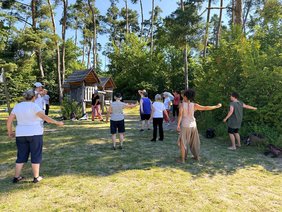 Gruppe von Erwachsenen macht gemeinsam sanfte Gymnastikübungen im Freien; sie stehen im Kreis auf einer Wiese und strecken die Arme aus. Im Hintergrund befinden sich Bäume und ein kleiner Holzunterstand, die Szene spielt bei sonnigem Wetter im Park.