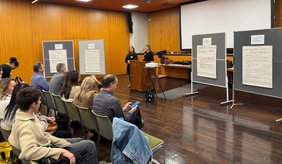 2 Frauen stehen am Rednerpult und vor ihnen viele Menschen auf Stühlen die zuhören.