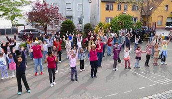 Eine große Gruppe aus Kindern und Senioren steht gemeinsam auf einem Hof und tanzt Line Dance. Viele heben die Arme in die Luft und bewegen sich synchron. Im Vordergrund tanzen Kinder und einige Erwachsene, im Hintergrund schauen weitere Teilnehmende zu. Die Stimmung wirkt fröhlich und gemeinschaftlich, umgeben von Wohnhäusern und Bäumen.