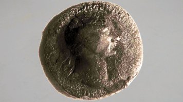 Das Foto zeigt eine stark gealterte, runde Münze vor hellem Hintergrund. Die Oberfläche wirkt rau und uneben, mit dunklen Verfärbungen und Ablagerungen, die Details teilweise überdecken. In der Mitte ist schemenhaft das Profil eines menschlichen Kopfes erkennbar, das nach rechts blickt. Die Konturen sind jedoch durch Abnutzung und Korrosion nur noch schwer auszumachen. Der Rand der Münze ist unregelmäßig. Es handelt sich bei der Münze um das älteste bekannte Geld Viernheims, geprägt unter dem römischen Kaiser Traian (98–117 n. Chr.).