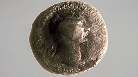 Das Foto zeigt eine stark gealterte, runde Münze vor hellem Hintergrund. Die Oberfläche wirkt rau und uneben, mit dunklen Verfärbungen und Ablagerungen, die Details teilweise überdecken. In der Mitte ist schemenhaft das Profil eines menschlichen Kopfes erkennbar, das nach rechts blickt. Die Konturen sind jedoch durch Abnutzung und Korrosion nur noch schwer auszumachen. Der Rand der Münze ist unregelmäßig. Es handelt sich bei der Münze um das älteste bekannte Geld Viernheims, geprägt unter dem römischen Kaiser Traian (98–117 n. Chr.).