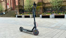E-Scooter mit der Apostelkirche und Blumenbeeten im Hintergrund. Pflasterboden der Innenstadt.
