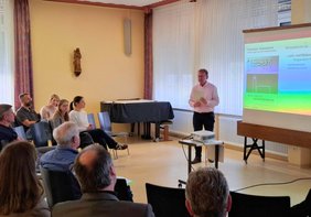 Bürgermeister Matthias Baaß steht vor einer Leinwand und spricht zu einer Gruppe von etwa 15 sitzenden Teilnehmenden in einem hellen Raum. Neben ihm ist eine Präsentation zu sehen, die sich mit Nutzungsmöglichkeiten von Kirchenstandorten befasst.