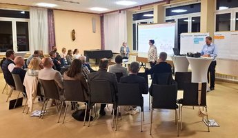 Mehrere Personen sitzen in Stuhlreihen und verfolgen aufmerksam eine Präsentation. Zwei Frauen stehen vorne im Raum und moderieren die Diskussion, während auf Stellwänden und einer Leinwand gesammelte Ideen und Notizen zu sehen sind.