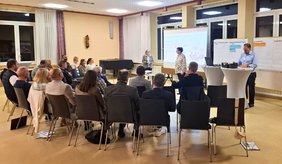 Mehrere Personen sitzen in Stuhlreihen und verfolgen aufmerksam eine Präsentation. Zwei Frauen stehen vorne im Raum und moderieren die Diskussion, während auf Stellwänden und einer Leinwand gesammelte Ideen und Notizen zu sehen sind.