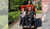 Zwei ältere Fahrgäste sitzen an einem sonnigen Tag in der orangefarbenen Viernheimer e-Rikscha und halten jeweils ein Eis in der Hand. Hinter ihnen steht ein lächelnder ehrenamtlicher Fahrer. Die Rikscha befindet sich auf einem Weg im Grünen, umgeben von Bäumen, Blumenbeeten und Häusern im Hintergrund. Die Szene vermittelt einen entspannten und fröhlichen Ausflug in angenehmer Atmosphäre.
