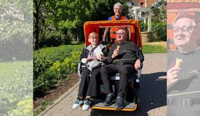 Zwei ältere Fahrgäste sitzen an einem sonnigen Tag in der orangefarbenen Viernheimer e-Rikscha und halten jeweils ein Eis in der Hand. Hinter ihnen steht ein lächelnder ehrenamtlicher Fahrer. Die Rikscha befindet sich auf einem Weg im Grünen, umgeben von Bäumen, Blumenbeeten und Häusern im Hintergrund. Die Szene vermittelt einen entspannten und fröhlichen Ausflug in angenehmer Atmosphäre.