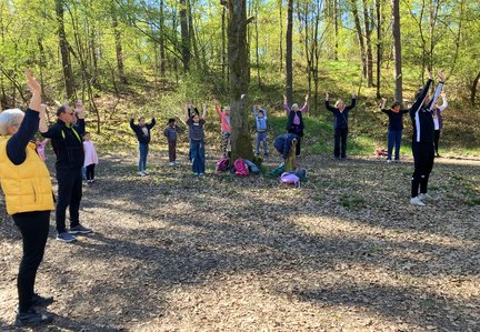 Eine Gruppe aus Kindern und älteren Menschen steht in einem Waldkreis auf einem mit Laub bedeckten Boden und macht gemeinsam Gymnastikübungen. Alle heben die Arme nach oben. In der Mitte liegen Rucksäcke am Fuß eines Baumes, umgeben von hohen Bäumen und frischem Grün.