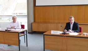 Bürgermeister Matthias Baaß und Amtsleiter Rudolf Haas (links) wenden sich im Rahmen einer Pressekonferenz an die Bürgerschaft in Viernheim. ©Stadt Viernheim Rudolf Haas und Bürgermeister Baaß sitzen je hinter einem Holztisch, auf dem Unterlagen liegen und ein Mikro steht