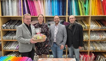 Vier Personen stehen in einem Fachgeschäft für Stoffe vor einem großen Regal, das farbenfrohe Stoffrollen sortiert nach Farben zeigt. Links im Bild hält Bürgermeister Matthias Baaß einen Geschenkkorb mit Zellophanverpackung. Neben ihm stehen Inhaberin Renate Müller, Alexander Schwarz und Claus Bunte von der Wirtschaftsförderung. Alle lächeln in die Kamera. Im Vordergrund liegen weitere bunte Stoffe und Nähzubehör.