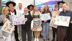 Gruppenfoto mit elf Personen bei der Vorstellung der „Vernemer Kerwe 2026“ in einem Innenraum. Einige Personen tragen traditionelle Kleidung mit schwarzen Hüten und roten Halstüchern. Im Mittelpunkt steht eine junge Frau im Dirndl, die ein großes Lebkuchenherz mit der Aufschrift „Vernemer Kerwe 2026“ hält. Weitere Personen präsentieren drei bunt gestaltete Kinderzeichnungen mit Kerwemotiven sowie Stofftaschen mit dem Aufdruck „Seit 1995“ und einer Illustration. Im Hintergrund ist auf einem Bildschirm ein farbenfrohes Motiv zur Vernemer Kerwe zu sehen. Die Gruppe steht dicht beieinander und blickt überwiegend lächelnd in die Kamera.