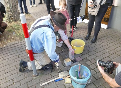 Der Künstler und Initiator des Stolpersteinprojekts Gunter Demnig kniet auf dem Gehweg und arbeitet an mehreren in das Pflaster eingelassenen Stolpersteinen. Um ihn herum stehen Arbeitsgeräte, Eimer und mehrere Zuschauende. Hinter ihm hält ein kleines Mädchen eine israelische Fahne.