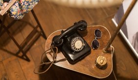 Auf einem kleinen Holztisch im Museum steht ein schwarzes Wählscheibentelefon. Daneben liegen eine Sonnenbrille und eine Getränkedose. Im Hintergrund sind ein gepolsterter Stuhl und weitere Teile der Ausstellung zu sehen. Das historische Telefon ist Teil der Dauerausstellung zur Geschichte Viernheims.