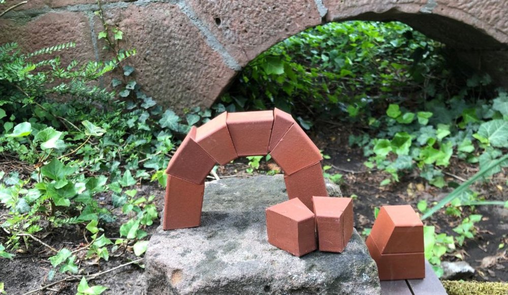 Kleine rote Bauklötze sind auf einem Stein zu einem Brückenbogen angeordnet. Daneben liegen weitere Bausteine. Im Hintergrund ist ein gemauerter Bogen zu sehen, umgeben von Efeu und anderen Pflanzen.