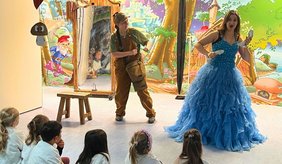 Zwei Darsteller der „Opernretter“ führen in der Kita Entdeckerland eine Kinderoper auf. Eine Frau im langen, hellblauen Märchenkleid und ein Mann in brauner Latzhose stehen vor einer bunten Kulisse mit Märchenwald und Schloss. Im Vordergrund sitzen mehrere Kinder auf dem Boden und verfolgen die Aufführung.