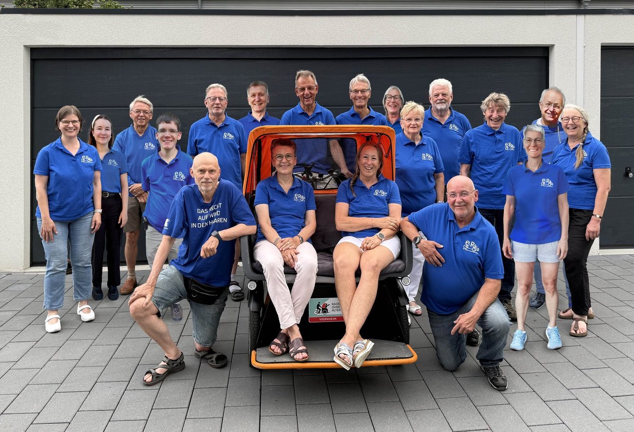 Vielen Personen stehen in blauen T-Shirts um die E-Rikscha