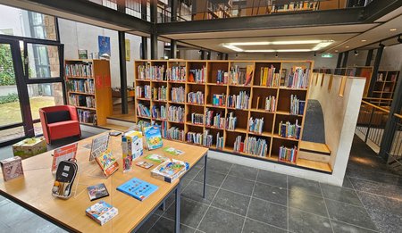 Innenansicht der Stadtbibliothek Viernheim mit gut gefüllten Bücherregalen, einem großen Tisch mit Büchern und Spielen sowie einem roten Sessel. Große Fenster geben den Blick auf einen begrünten Innenhof frei.