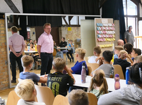 Bürgermeister Matthias Baaß steht im Raum und spricht zu einer Gruppe von Kindern, die ihm aufmerksam zuhören. Er trägt ein rosafarbenes Hemd und dunkle Jeans. Hinter ihm ist ein großer Spiegel, in dem sein Rücken reflektiert wird. Die Kinder sitzen an Tischen und schauen ihn an. Im Hintergrund sieht man weitere Jugendliche und Erwachsene. Rechts im Bild hängt eine Pinnwand mit vielen bunten Notizzetteln.