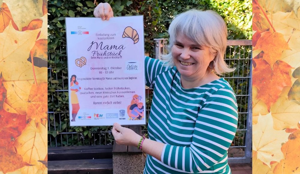 Eine lächelnde Frau hält im Freien ein Plakat für ein kostenloses „Mama Frühstück“ hoch. Darauf wird zu einem gemütlichen Vormittag für Mamas und ihre kleinen Begleiter eingeladen. Das Foto ist seitlich mit herbstlich gefärbten Blättern eingerahmt.