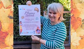 Eine lächelnde Frau hält im Freien ein Plakat für ein kostenloses „Mama Frühstück“ hoch. Darauf wird zu einem gemütlichen Vormittag für Mamas und ihre kleinen Begleiter eingeladen. Das Foto ist seitlich mit herbstlich gefärbten Blättern eingerahmt.
