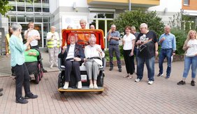 Zwei Frauen und viele Menschen dahinter sitzen auf einer orangenen Rikscha.