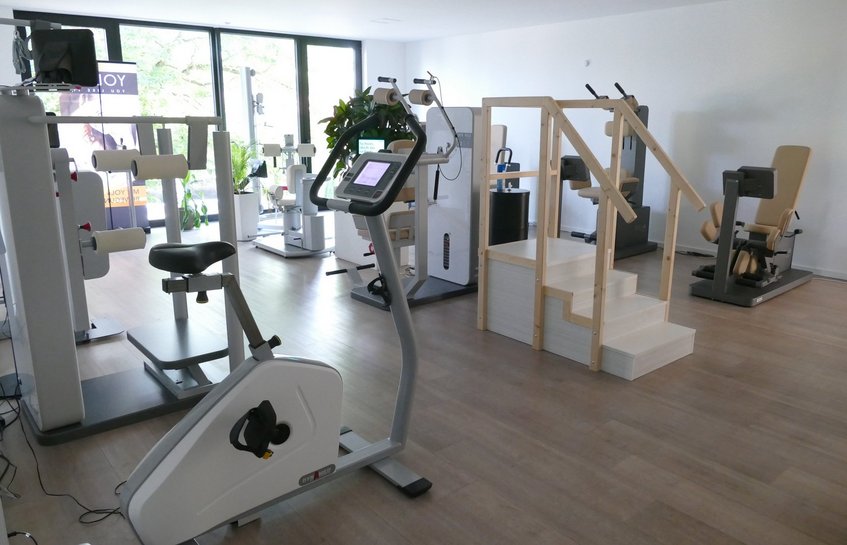 Ein heller, modern eingerichteter Trainingsraum für die Neurorehabilitation mit großen Fenstern und hellem Holzboden. Im Raum stehen mehrere medizinische Trainingsgeräte in Grau und Weiß. Im Vordergrund befindet sich ein Fahrradergometer mit Bildschirm. Rechts daneben steht eine kleine, dreistufige Übungstreppe aus hellem Holz mit beidseitigen Handläufen. Im Hintergrund sind weitere Geräte mit Sitzen, Griffen und Polstern zu sehen. Einige Grünpflanzen stehen vor den Fenstern. Personen sind nicht zu sehen.
