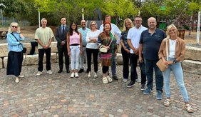 Eine Gruppe von zwölf Personen steht für ein gemeinsames Foto auf einem gepflasterten Platz. Im Hintergrund befinden sich Bäume, Grünflächen und Spielgeräte eines Spielplatzes.