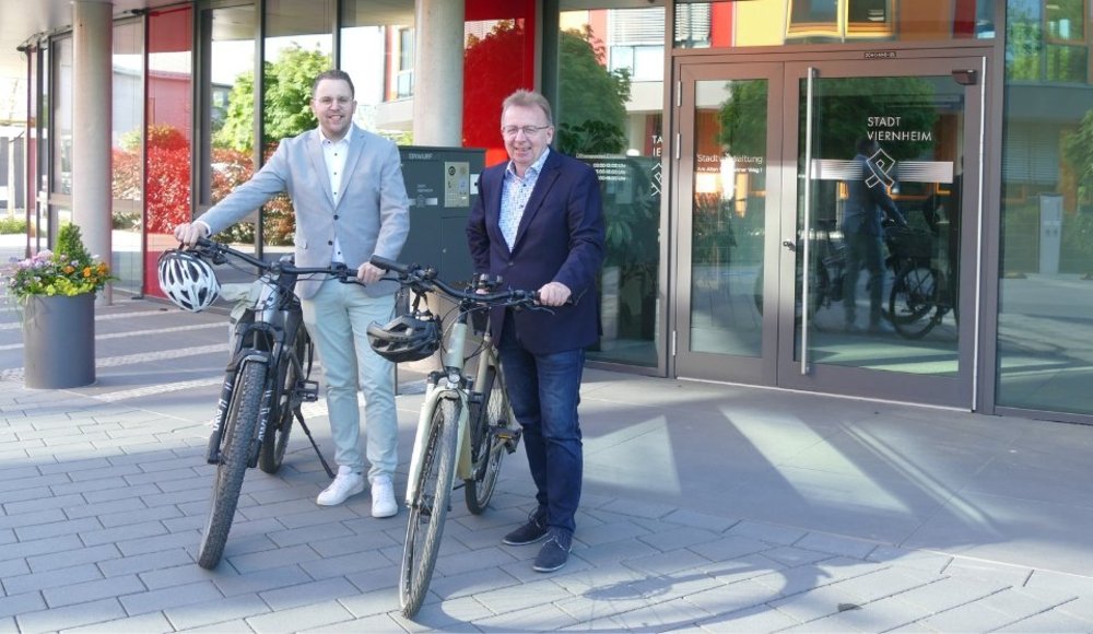 Das Foto zeigt Bürgermeister Matthias Baaß und Ersten Stadtrat Jörg Scheidel, die jeweils neben einem Fahrrad vor dem Eingangsbereich des neuen Rathauses stehen. Beide lächeln freundlich in die Kamera. Bürgermeister Baaß trägt eine dunkle Jacke, ein Hemd und Jeans. Erster Stadtrat Scheidel ist heller gekleidet, mit einem Sakko, Hemd und hellen Hosen. An den Fahrrädern ist auch jeweils ein Fahrradhelm zu sehen. Die Fahrräder stehen auf einem gepflasterten Vorplatz. Im Hintergrund befindet sich eine Glasfassade mit Eingangstüren, auf denen der Schriftzug „Stadt Viernheim“ zu erkennen ist. Durch die Glasflächen spiegeln sich die Umgebung und Teile des Gebäudes. Pflanzkübel und moderne Architektur unterstreichen den modernen Charakter der Szene. Das Bild vermittelt den Eindruck eines offiziellen Termins im Freien und verweist auf eine Aktion rund ums Fahrradfahren.