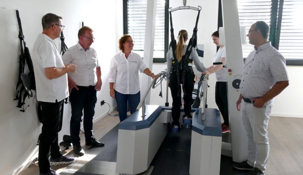 In einem hellen Therapieraum steht eine Person mit einem schwarzen Gurtsystem gesichert auf einem Laufband zur Gangrehabilitation. Eine Therapeutin und ein Therapeut begleiten die Übung. Drei weitere Männer stehen daneben und beobachten die Vorführung. Das Gerät verfügt über seitliche Haltegriffe und eine Aufhängung zur Unterstützung der Bewegungen.
