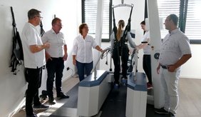 In einem hellen Therapieraum steht eine Person mit einem schwarzen Gurtsystem gesichert auf einem Laufband zur Gangrehabilitation. Eine Therapeutin und ein Therapeut begleiten die Übung. Drei weitere Männer stehen daneben und beobachten die Vorführung. Das Gerät verfügt über seitliche Haltegriffe und eine Aufhängung zur Unterstützung der Bewegungen.