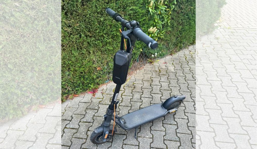 Ein schwarzer E-Scooter steht auf einem gepflasterten Weg vor einer dichten grünen Hecke. Der Roller ist schräg von vorne fotografiert und verfügt über einen breiten Lenker, eine schwarze Tasche an der Lenkstange, ein rutschfestes Trittbrett sowie Schutzbleche über den Rädern. Ein orangefarbenes Kabel verläuft entlang der Lenkstange zum Vorderrad. Links und rechts wird das hochformatige Foto durch aufgehellte Ausschnitte des Hintergrunds ergänzt.