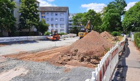 Blick auf eine Straßenbaustelle in der Saarlandstraße. Die Fahrbahn ist großflächig geöffnet und mit einer Schotterschicht vorbereitet. Im Vordergrund liegen mehrere große Erdhaufen, dahinter arbeiten ein Bagger und eine Walze auf der Baustelle. Weiße Absperrbaken begrenzen den Arbeitsbereich. Rechts verläuft ein Gehweg entlang der Baustelle. Im Hintergrund stehen mehrgeschossige Wohnhäuser und große, grüne Bäume bei sonnigem Wetter.