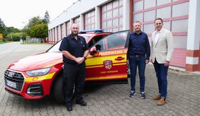 Drei Männer stehen nebeneinander vor dem neuen Kommandowagen der Feuerwehr. Von rechts nach links: Erster Stadtrat Jörg Scheidel in Anzug und Krawatte, daneben Tobias Kettner und Cornelius Hisge, beide in Feuerwehruniform. Im Hintergrund ist das rot-weiße Einsatzfahrzeug mit geöffneter Seitentür zu sehen.