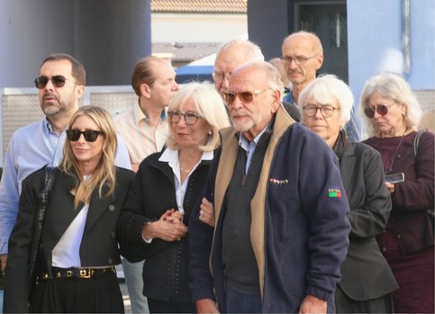 Eine Gruppe von Erwachsenen steht dicht beieinander im Freien und hört aufmerksam zu. In der Bildmitte steht Susanne Newmark, die Tochter von Frieda Horn, neben ihrem Mann. Mehrere Personen tragen Sonnen- oder normale Brillen.