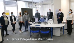 Matthias Baaß (2.v.r) und Büroleiter Jörg Gutperle (3.v.r.) stellen bei einem Pressetermin den erweiterten Service im Bürgerbüro vor. ©Stadt Viernheim Mitarbeitende des Bürgerbüros, Bürgermeister Matthias Baaß und Büroleiter Jörg Gutperle stehen im Halbkreis mit Masken, eine Person sitzt am Schreibtisch