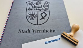 Graue Urkundenmappe der Stadt Viernheim mit Stadtwappen. Im Vordergrund liegen ein weißes Blatt mit dem Aufdruck „Standesamt“ und ein entsprechender Holzstempel. Bei der Abteilung Standesamt der Stadt Viernheim ist derzeit mit längeren Wartezeiten zu rechnen.
